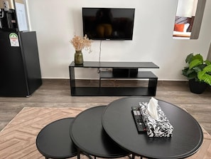 Un salon moderne avec un téléviseur, un meuble de rangement noir, deux petites tables rondes noires, un tapis beige et une plante en pot.