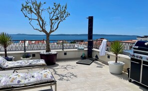 Een terras op het dak met uitzicht op zee, een barbecue, lounge fauteuils en potplanten.