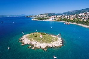An aerial view of a coastal town with clear blue waters, a rocky island with a lighthouse, and a densely built urban area in the background. - Modern apartment with sea view and balcony in Novi Vinodolski (Novi Vinodolski)