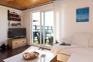 Un salon doté d’un téléviseur à écran plat, d’un canapé blanc, d’une table basse avec un saladier, et d’une porte-fenêtre coulissante donnant sur un espace extérieur aménagé avec des meubles en osier.