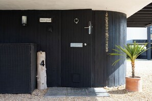 Une porte en bois noire avec une pancarte indiquant « MAISON DE PLAGE », une plaque portant le numéro « 4 » et un pot de fleurs placé à côté.