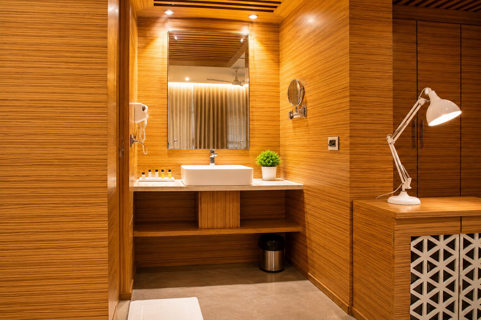 A modern bathroom with wooden paneling, a white sink, a mirror, a plant, a trash bin, and a white desk lamp.