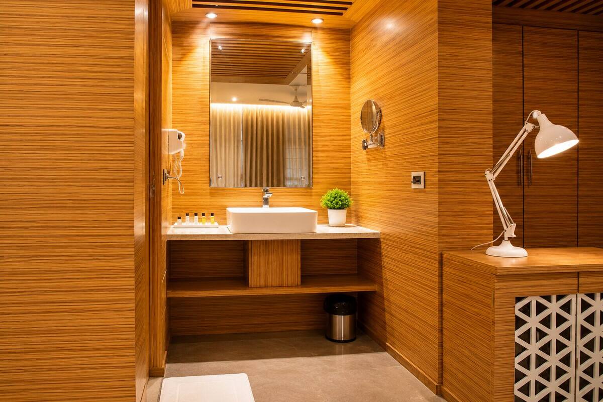 Un baño moderno con paneles de madera, un lavabo blanco, un espejo, una planta, un cubo de basura y una lámpara de escritorio blanca.