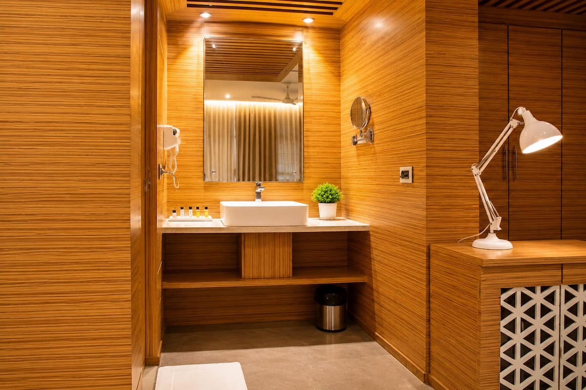 A modern bathroom with wooden paneling, a white sink, a mirror, a plant, a trash bin, and a white desk lamp.
