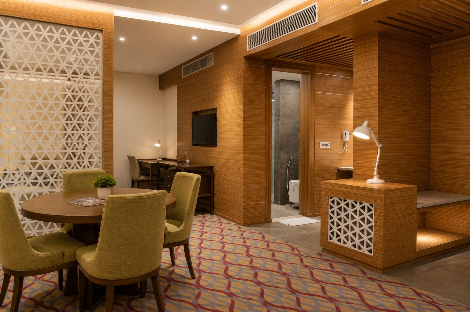 A modern hotel room with a wooden accent wall, a desk with a lamp, a small seating area, and a patterned carpet.