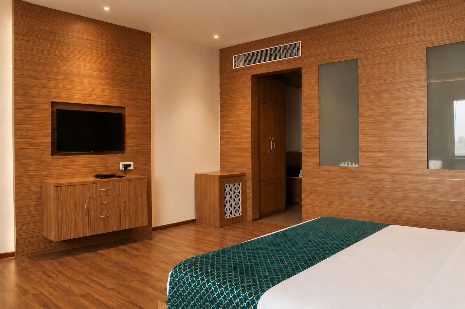 A modern bedroom with a wooden wall panel, a TV mounted above a wooden cabinet, a bed with a patterned bedspread, and a door leading to a bathroom.