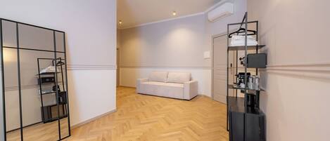 A hotel room with a herringbone floor, a white sofa, a mirrored wardrobe, and a black shelf unit with a coffee machine.