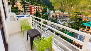 Ein Balkon mit zwei grünen Stühlen und einem kleinen Tisch, mit Blick auf eine gebirgige Landschaft und Gebäude.