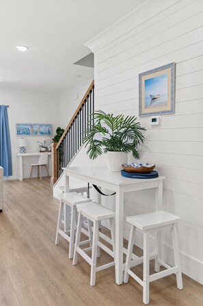 A modern kitchen with white cabinetry, a wooden staircase, and a dining area with white stools. - Coastal Chic Steps to Beach Pet Friendly (Myrtle Beach)