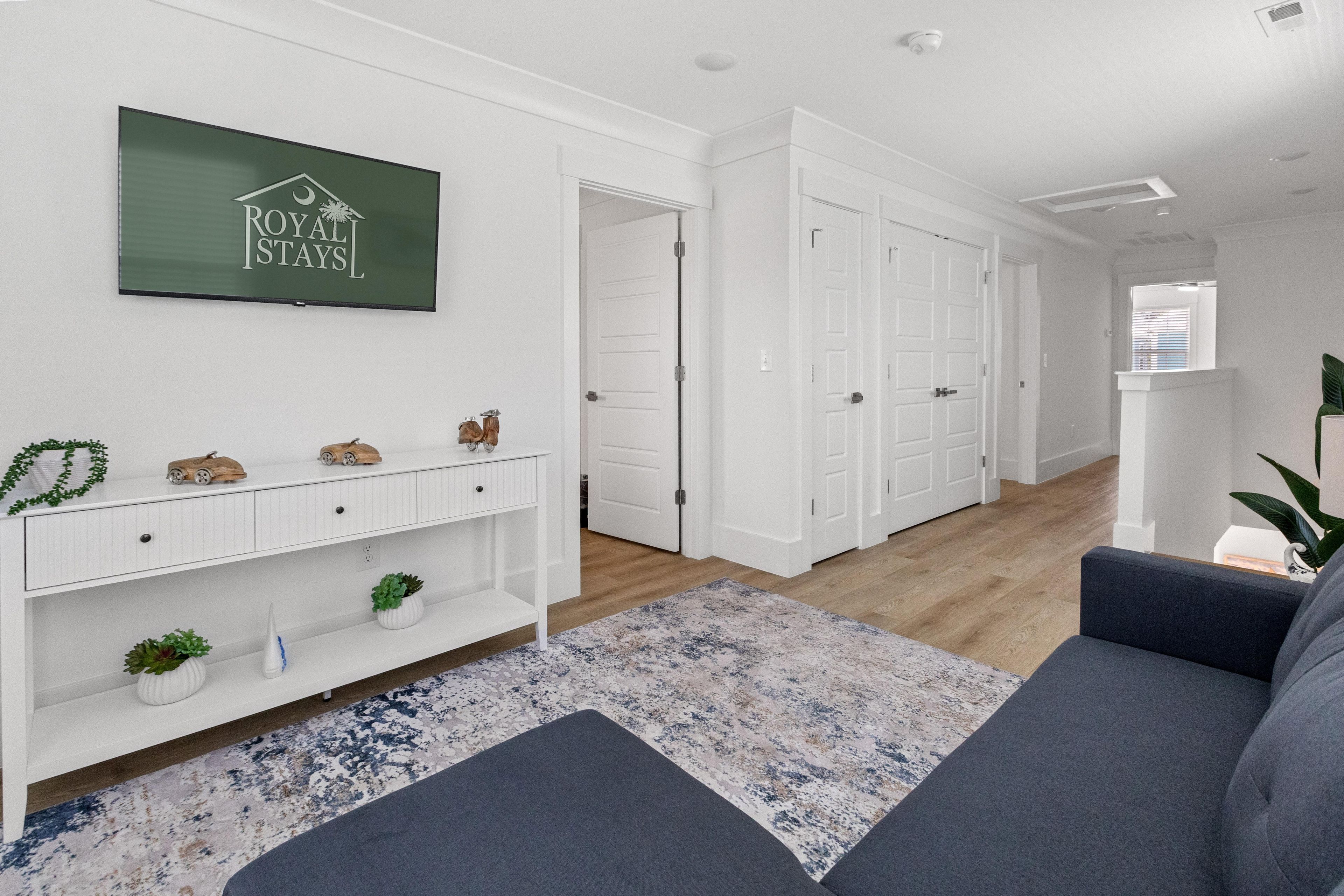 A modern living room with a grey sofa, a mounted television displaying 'ROYAL STAYS', and a hallway leading to a door.