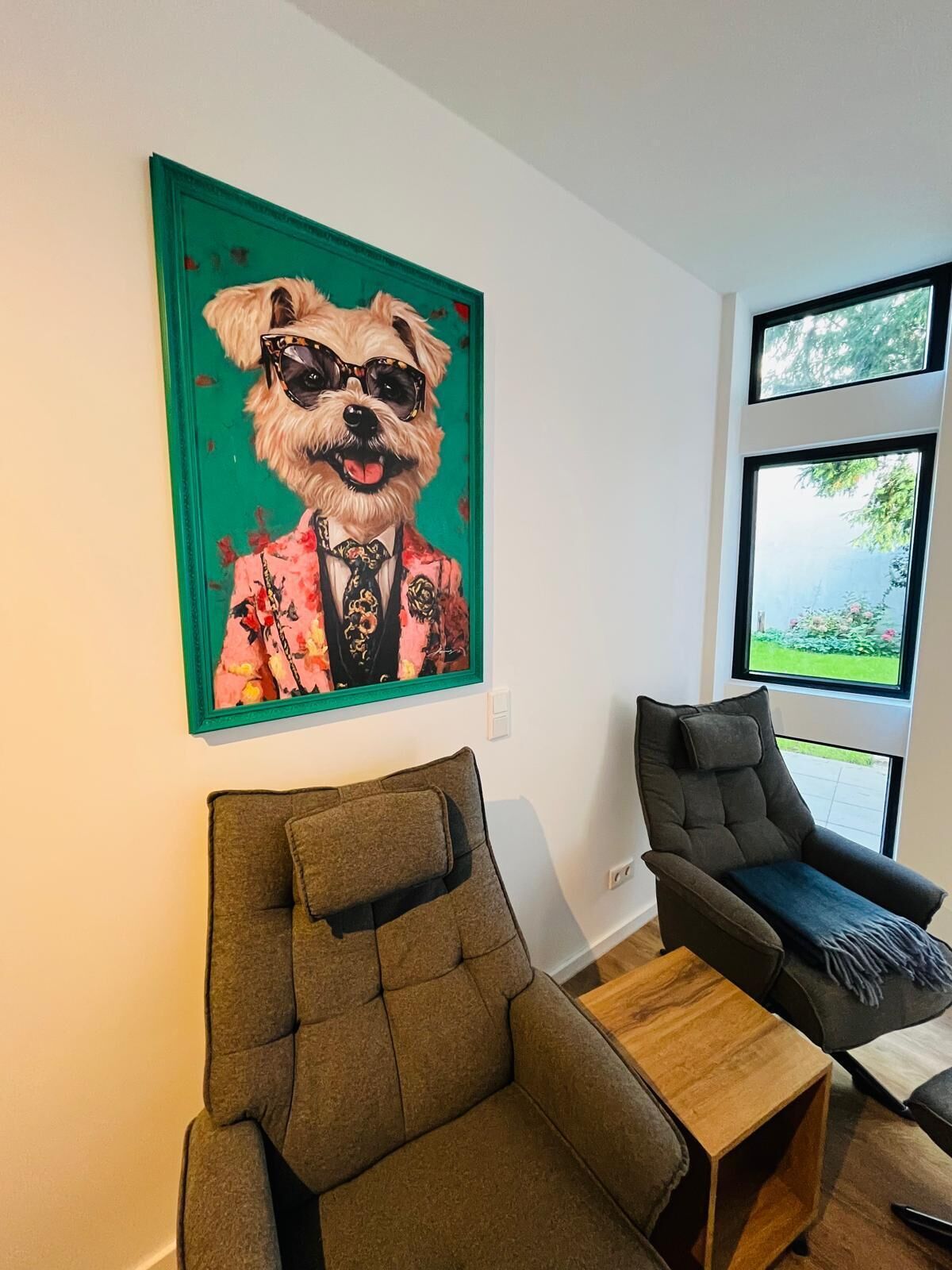 Un salón moderno con un cuadro grande de un perro trajeado, un sillón gris, una mesita de madera y una ventana con vistas a zonas verdes.