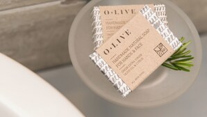 Una pastilla de jabón con etiqueta que indica que es jabón natural artesanal para manos y rostro, con aceite de oliva griego extra virgen y cítricos.