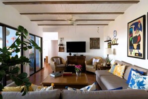 A living room with a sofa, coffee table, and television. There are wicker chairs, a plant, and artwork on the walls.
