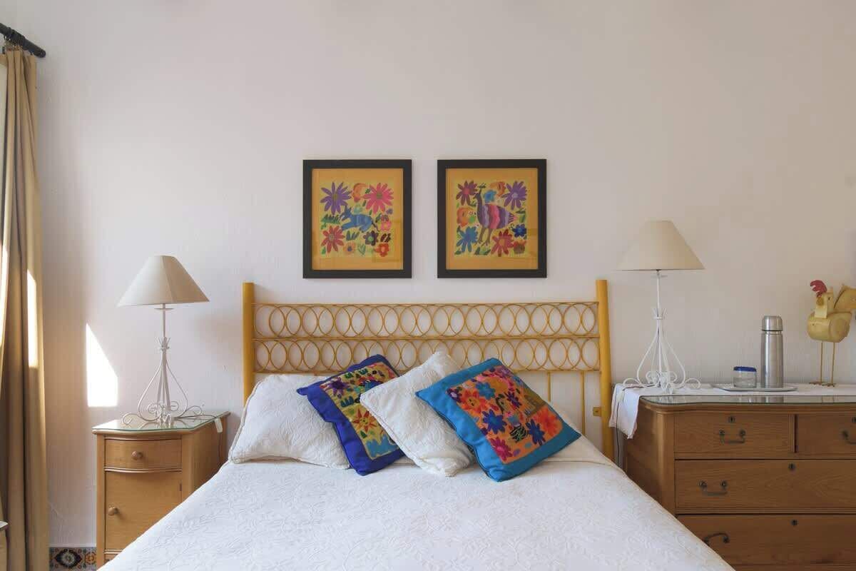 A neatly arranged bedroom with a bed, two pillows, a bedside table with a lamp, a dresser, and two framed artworks on the wall.