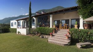 Una casa de dos pisos con un patio, escalones que conducen a la entrada y montañas al fondo.