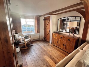 A room with wooden flooring, a rocking chair, a sofa, a wooden cabinet, a large window, and a mirrored wall. - 3 bedroom cottage with garden in Arundel (West Sussex)