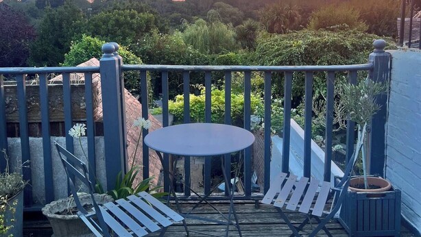 Una terraza de madera con una mesa redonda y dos sillas, rodeada de vegetación y una barandilla.