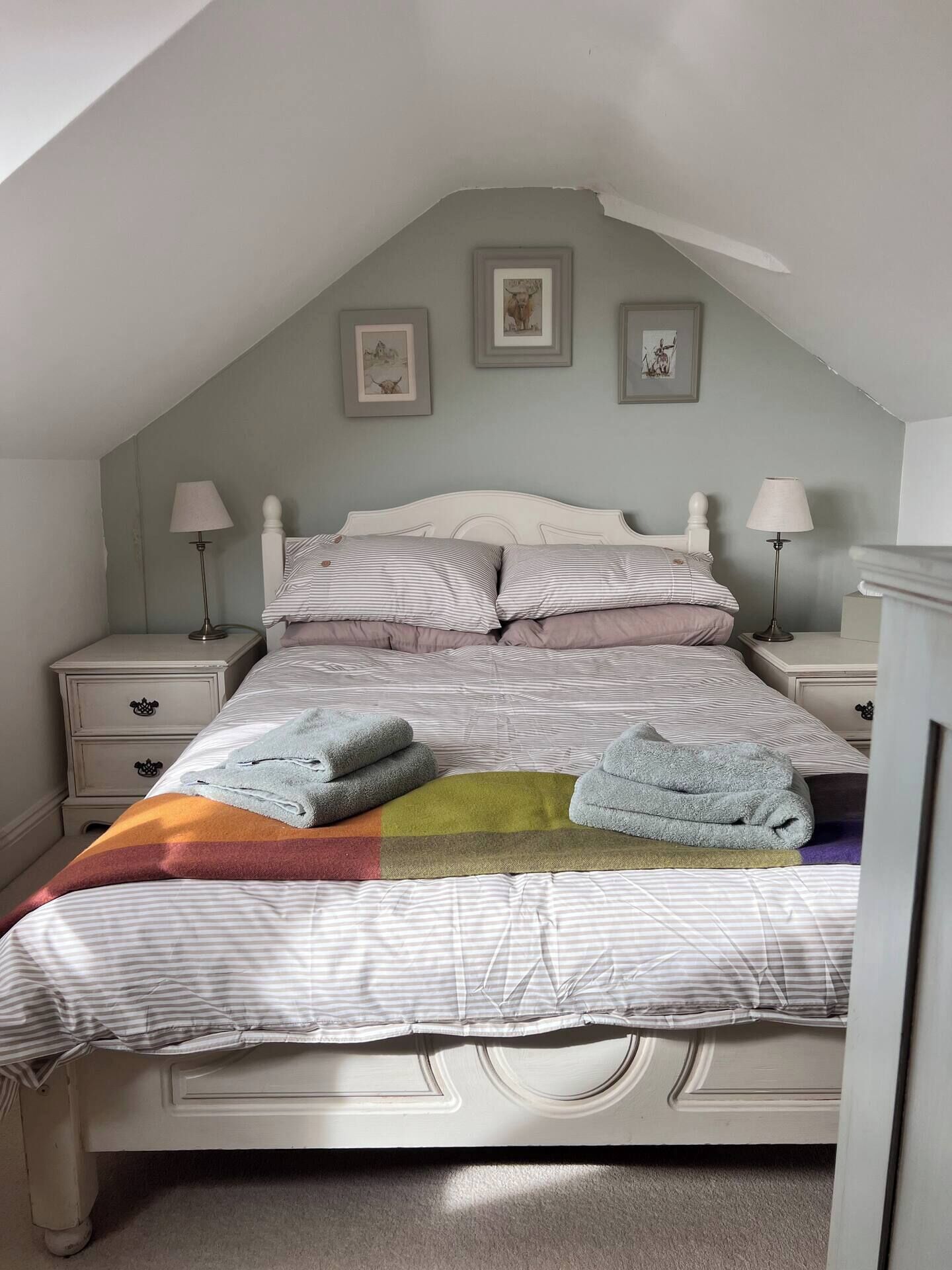 Un dormitorio con techo inclinado, una cama con ropa de cama a rayas, dos lámparas de noche y una manta de colores.
