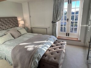 Een slaapkamer met een groot bed, een nachtkastje, een radiator, een raam met gordijnen en een deur.
