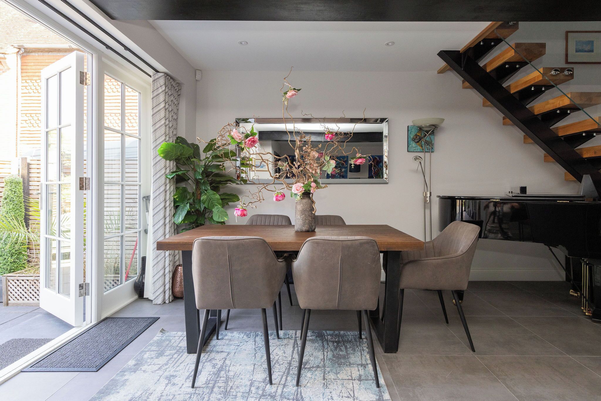 A dining area with a wooden table, six chairs, a vase with flowers, a large mirror, and a staircase.