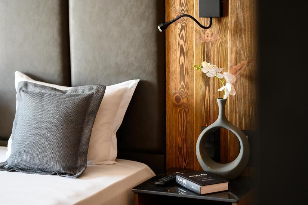 Una cama bien hecha con almohadas, una mesita de noche con un libro y un control remoto, y una pared de madera con un objeto circular.