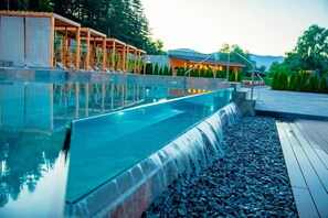 Una piscina con cascada, rodeada por una terraza de madera y un edificio con asientos al aire libre.