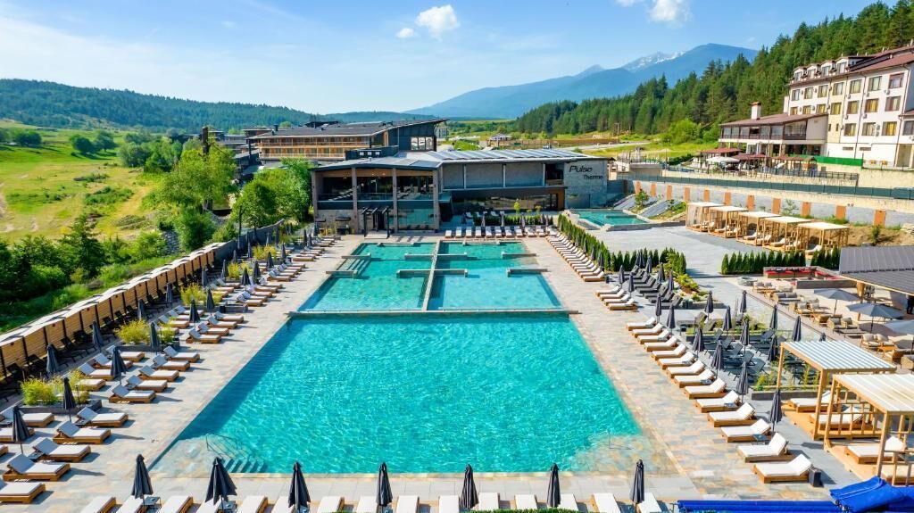 A hotel with a large outdoor swimming pool, lounge areas with sunbeds and umbrellas, and a mountainous backdrop.