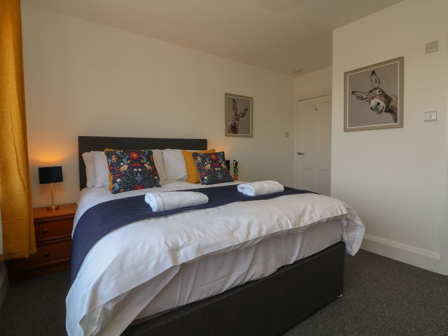 Una cama bien hecha con un cabecero oscuro, ropa de cama blanca y almohadas decorativas. Hay una mesita de noche con una lámpara y un cuadro colgado en la pared.