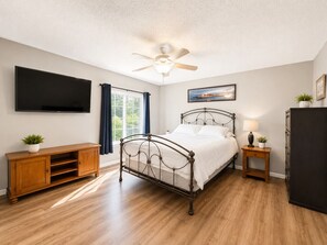 Ein Schlafzimmer mit einem Bett, einem Deckenventilator, einem Fernseher, einem Holzschrank, einem Nachttisch und einem Fenster mit Vorhängen.