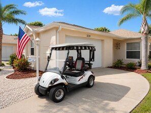 Ein Golfwagen vor einem Haus mit einer Garage, auf der die Nummer 2120 steht, einer amerikanischen Flagge und einem kleinen Busch mit roten Blüten.