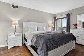 A bedroom with a bed, two bedside tables, a dresser, a window with curtains, and a view. - Ocean One 215 - 2BR/2BA Direct Oceanfront (Hilton Head Island)