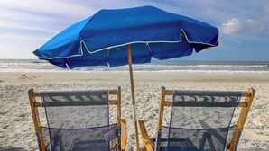 Zwei Liegestühle mit einem blauen Sonnenschirm am sandigen Strand.