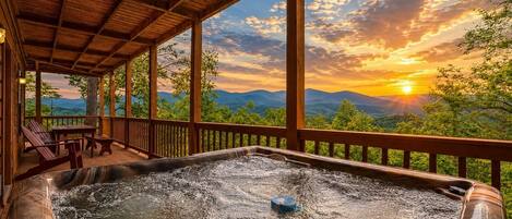 A hot tub with a wooden deck, a table, and chairs, overlooking a mountainous landscape during sunset.