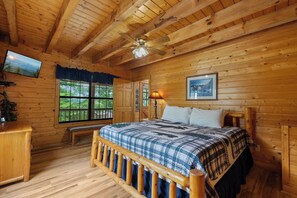Ein Schlafzimmer einer Holzhütte mit einem Bett, einer Bank, einem Schreibtisch, einem Deckenventilator und einem an der Wand befestigten Fernseher.