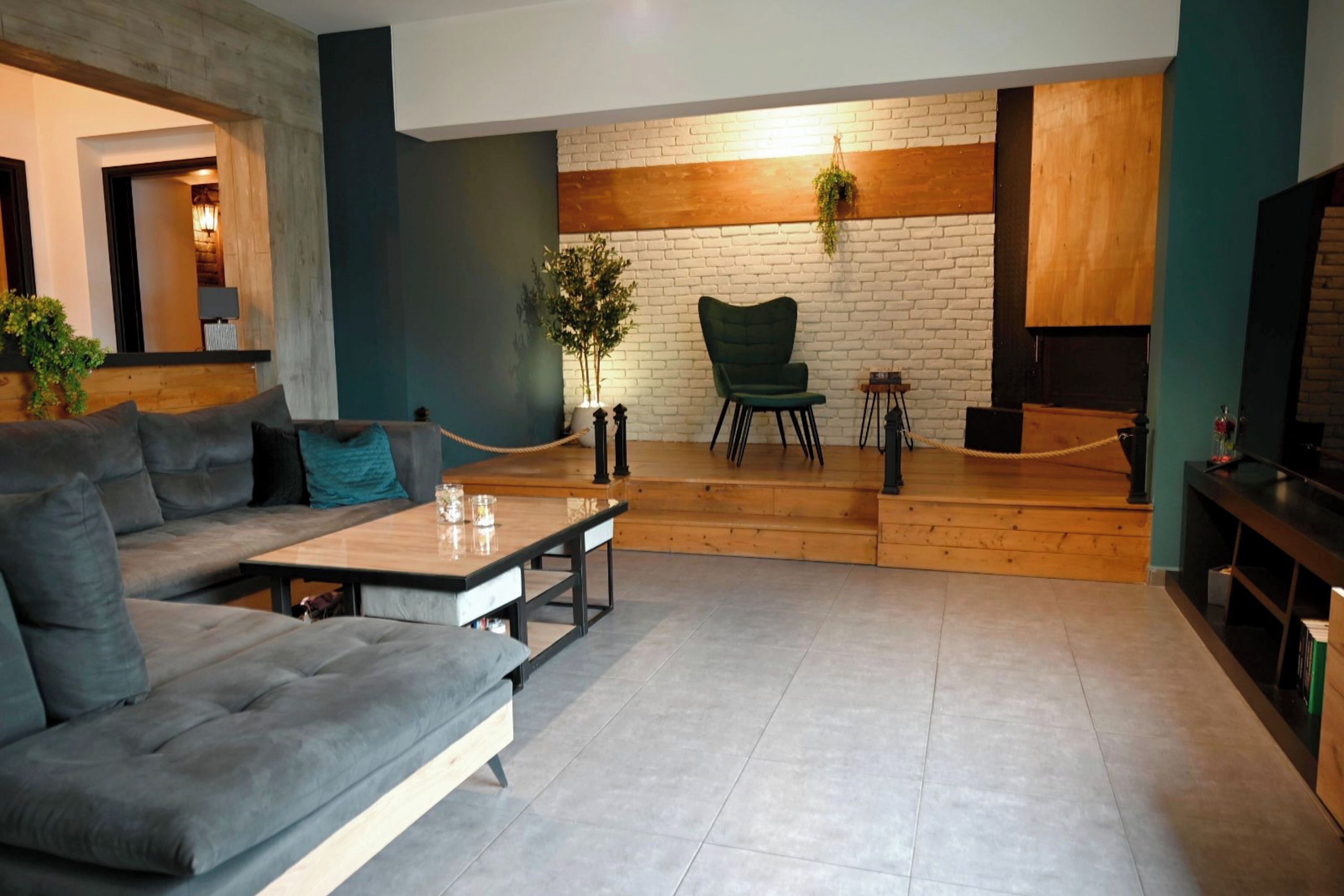 A modern living room with a sofa, a wooden coffee table, a green armchair, a brick wall, and a hanging plant.