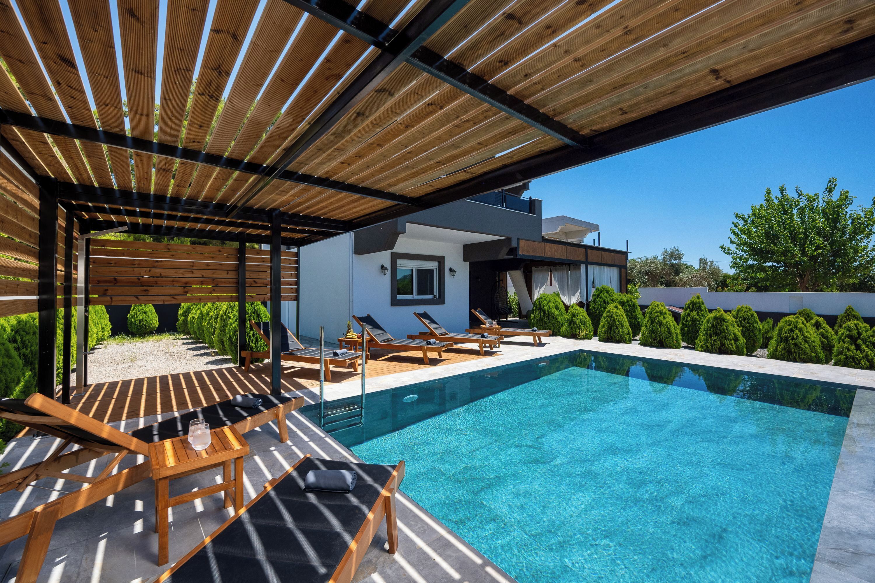 A modern outdoor pool area with wooden loungers, a clear pool, and a covered seating area.