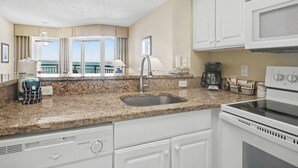 A kitchen with granite countertops, a stainless steel sink, a built-in oven, white cabinetry, and a view of the ocean through large windows. - Oceanview 1BR Suite + Lazy River & Hot Tub (Myrtle Beach)