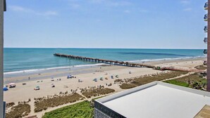 A beach view with a pier extending into the ocean, a sandy beach with umbrellas, and a building with a flat roof in the foreground. - Oceanview 1BR Suite + Lazy River & Hot Tub (Myrtle Beach)