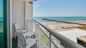 A balcony with white plastic chairs, a view of a beach, and a pier extending into the water. - Oceanview 1BR Suite + Lazy River & Hot Tub (Myrtle Beach)