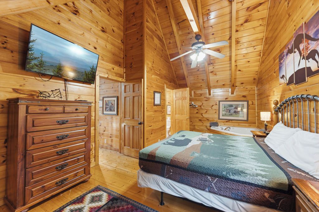 Interior de una cabaña de madera con una cama, cómoda, televisor, ventilador de techo y cuadros enmarcados.