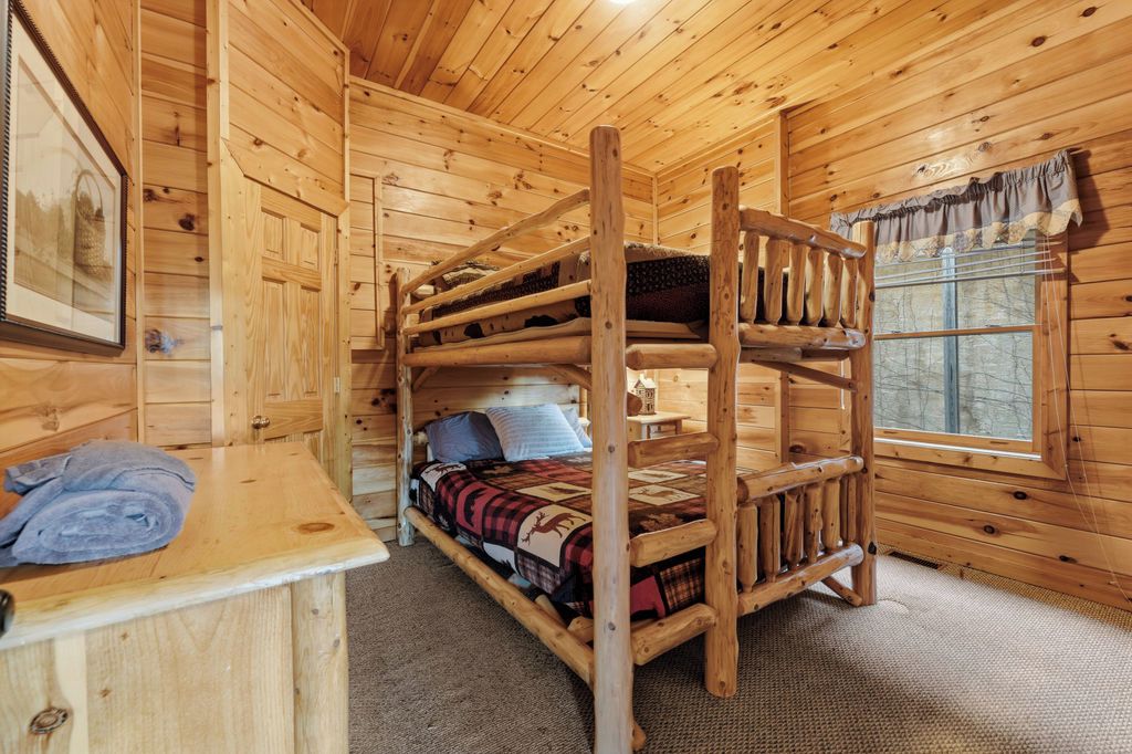A wooden cabin room with bunk beds, a small desk, and a window with curtains.