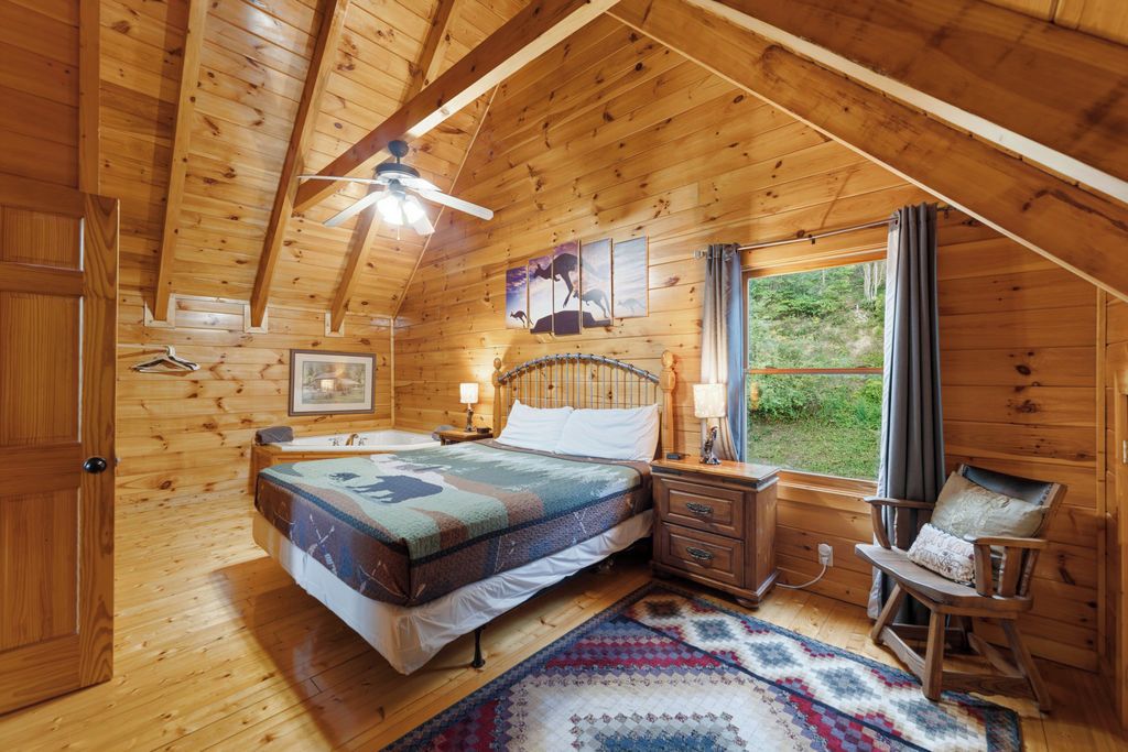Une chambre dans une cabane en bois, avec un lit, une table de chevet, une lampe, une chaise et une fenêtre avec des rideaux.