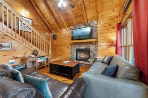 A log cabin interior with a stone fireplace, a television mounted above it, a staircase with wooden railings, and a leather sofa. - Amazing Rocky Top Views: Vistas That Wow (Pigeon Forge)