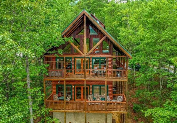 A multi-story log cabin with a balcony on the second floor, surrounded by dense forest. - Forget Me Not | Theater | Hot Tub | Mt LeConte Views | Sleeps 18 (Pigeon Forge)