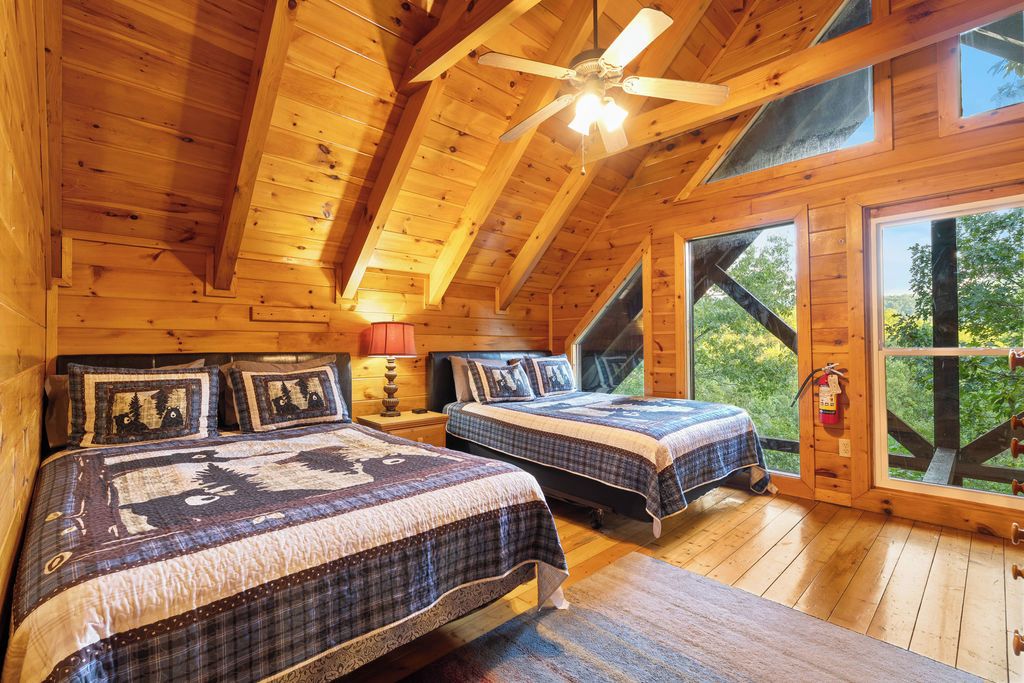A wooden cabin bedroom with two beds, a ceiling fan, and large windows.