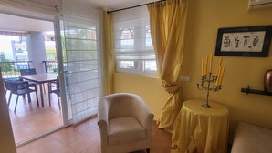 Una sala de estar con sofá, sillón y una pequeña mesa con un candelabro. Hay una puerta corrediza de vidrio que da a un área exterior con mesa y sillas.