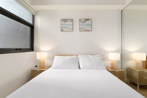 Quarto de hotel com uma cama, duas mesas de cabeceira com luminárias, uma janela e duas imagens emolduradas na parede.