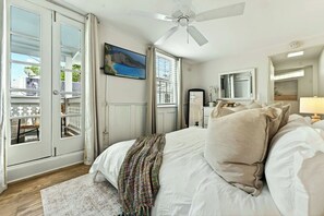 Ein Schlafzimmer mit einem Bett, einem Deckenventilator, einem Fenster mit Jalousien, einer Tür, die auf einen Balkon führt, und einem Gemälde an der Wand.