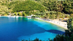 Ett kustområde med klart, blått vatten, en sandstrand och ett kuperat landskap i bakgrunden.