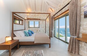 Ein Schlafzimmer mit Steinwand, einem Himmelbett, einem Nachttisch und einem Vorhang. Großes Fenster mit Blick.